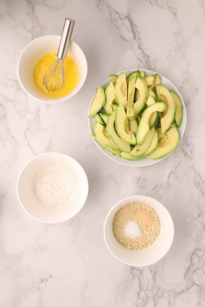 Avocado slices and three bowls with egg, flour, and seasoning breadcrumb mix to dip them in