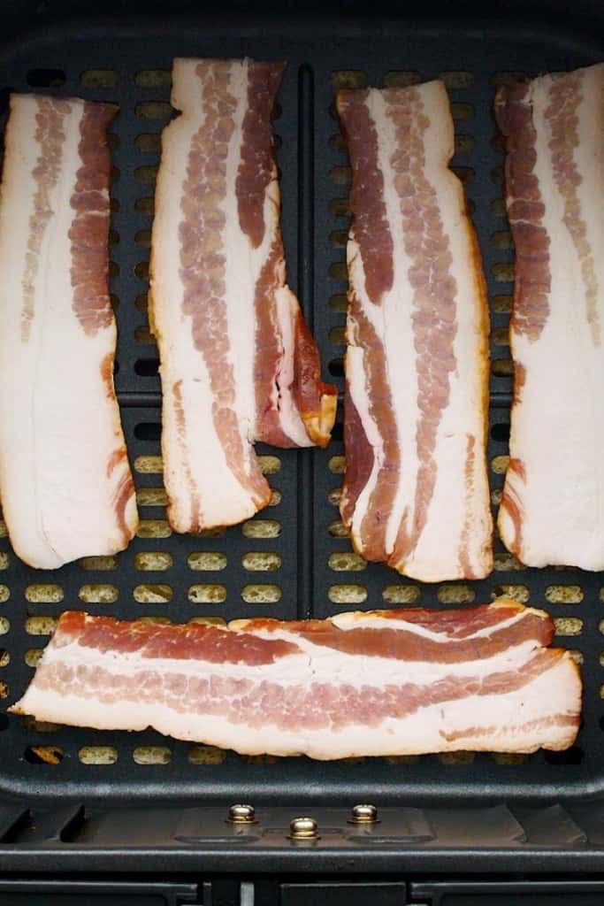 Raw bacon in air fryer basket with a piece of bread under the basket