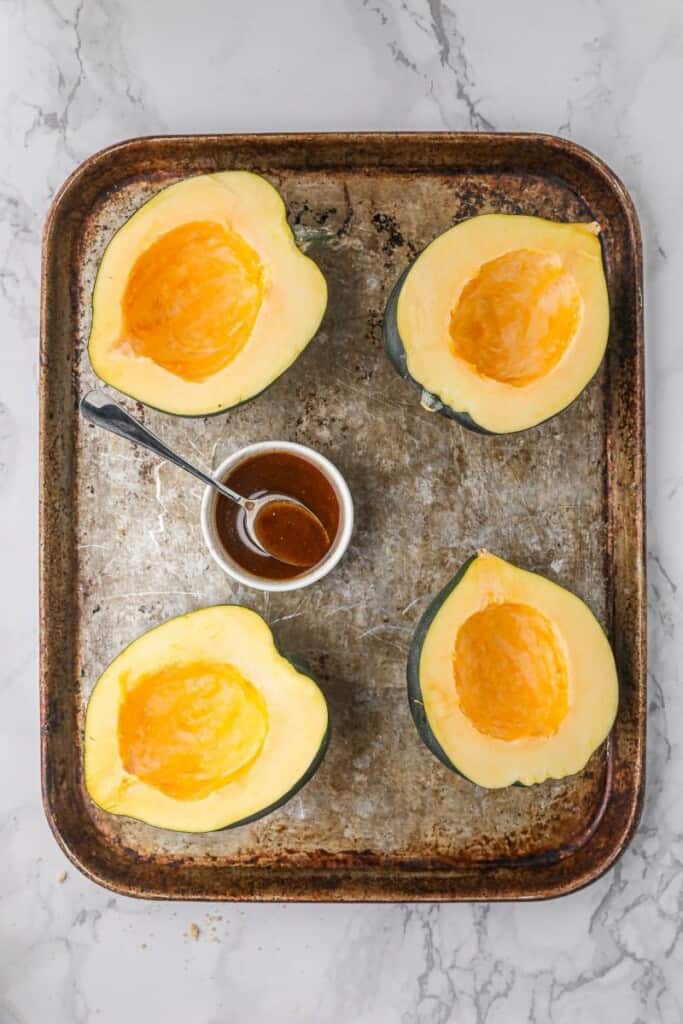 Raw acorn squash on a pan with the brown sugar butter mixture in a bowl with a spoon