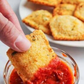 Hand dipping fried ravioli in a bowl of marinara sauce with fried ravioli in the background on a white plate