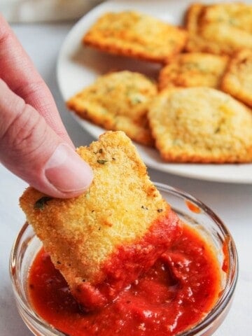 Hand dipping fried ravioli in a bowl of marinara sauce with fried ravioli in the background on a white plate