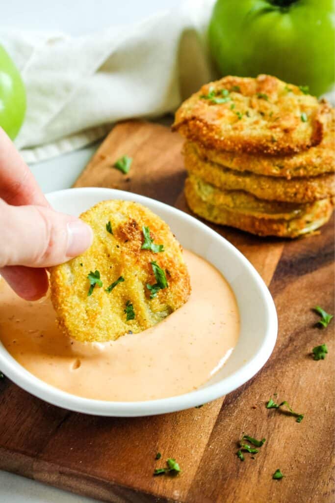 air fryer fried green tomato being dipped in a spicy mayo sauce with more slices behind it.