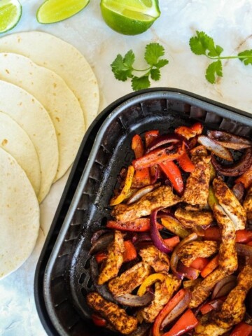 Cooked chicken fajitas inside air fryer with tortillas and lime wedges next to it