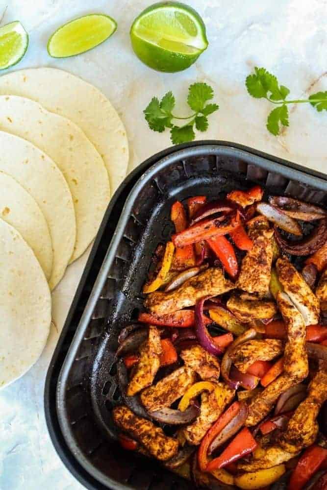 Cooked chicken fajitas inside air fryer with tortillas and lime wedges next to it