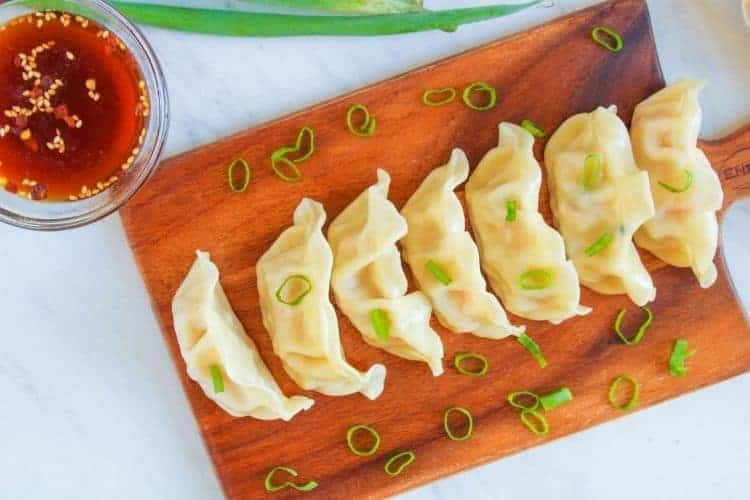 Dumplings on cutting board with green onions on top with bowl of sauce in the corner