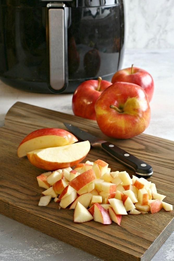 Chopped apples on a cutting board with full apples, a knife, and air fryer in background