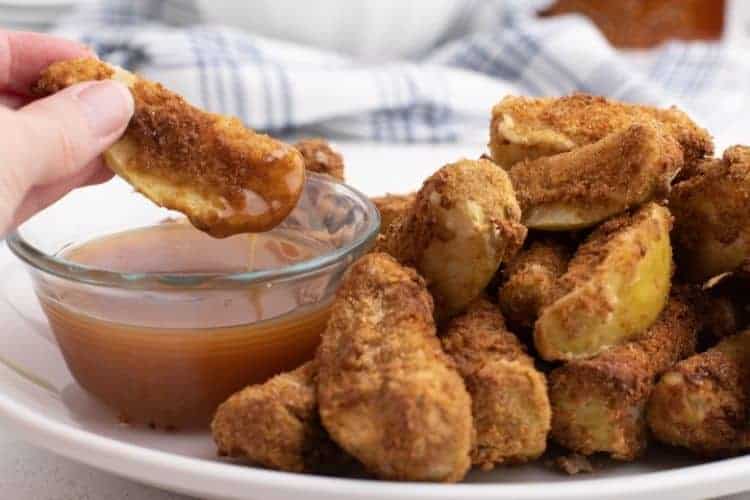 Air fryer apples on a white plate with one wedge being dipped in caramel