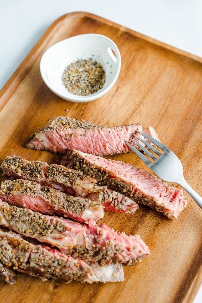 Sliced Air Fryer Ribeye Steak on a cutting board with a fork and a bowl of seasoning on the side