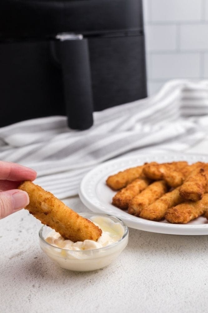 Fish Sticks being dipped into tartar sauce with plate of fish sticks and air fryer in the back