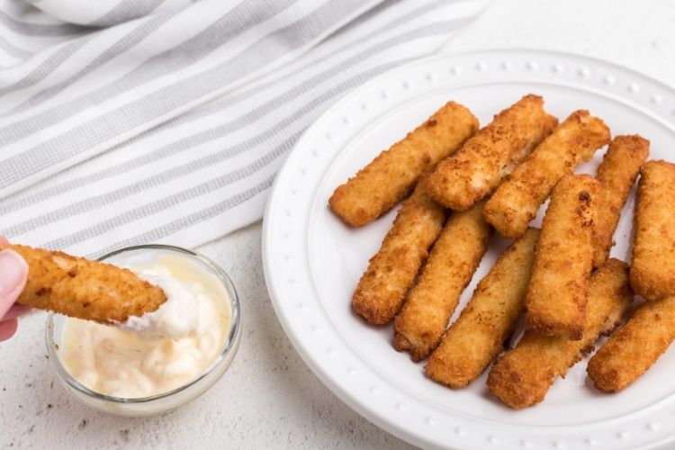 Fish stick being dipped into tartar sauce with plate of fish sticks to side