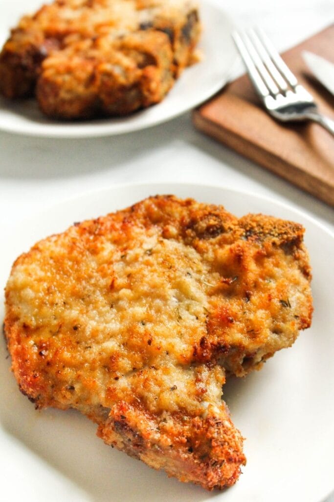 Air Fryer Bone-in Breaded Pork Chop on a white plate with fork and knife peaking out of the corner of picture