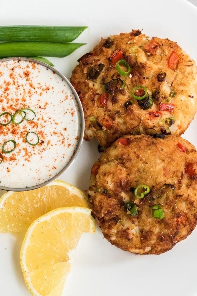 Two air fryer crab cakes on a white plate with dipping sauce and lemon wedges