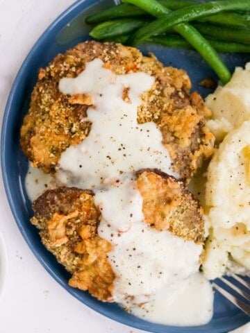 Air Fryer Chicken Fried Steak covered in gravy on a blue round plate with green beand and mashed potatoes