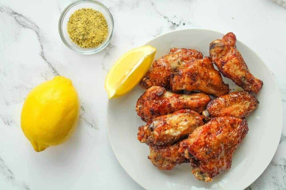 Air Fryer Lemon Pepper Chicken Wings on a white plate with a lemon slice and full lemon and small bowl of lemon pepper seasoning on side