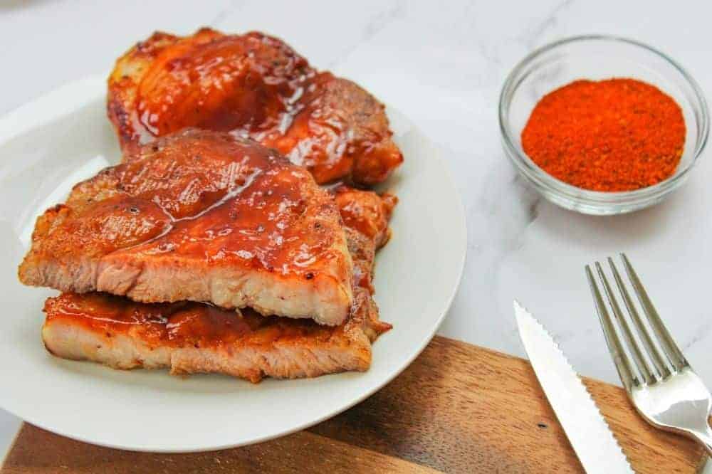 Air Fryer Pork Steaks with BBQ sauce on a white plate with small bowl of BBQ dry rub and fork and knife peaking out of the corner