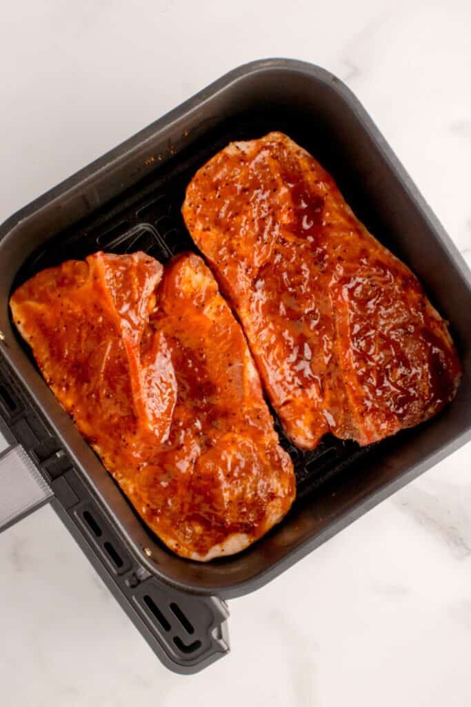 Raw pork steaks in an air fryer basket ready to be cooked with BBQ sauce on them.