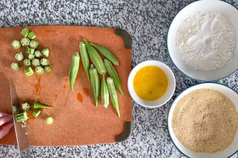 Cutting okra into pieces on a bown cutting board with 3 bowls of ingredients to the side