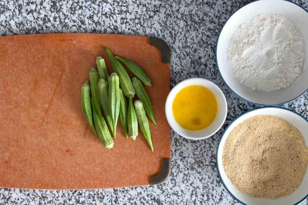 Full okra on a cutting board with bowls of egg, flour, and breadcrumb mixture