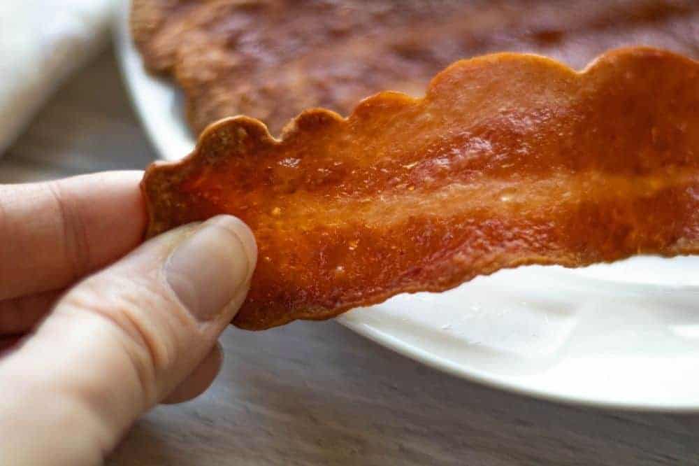 Hand holding a piece of turkey bacon with white plate in the background