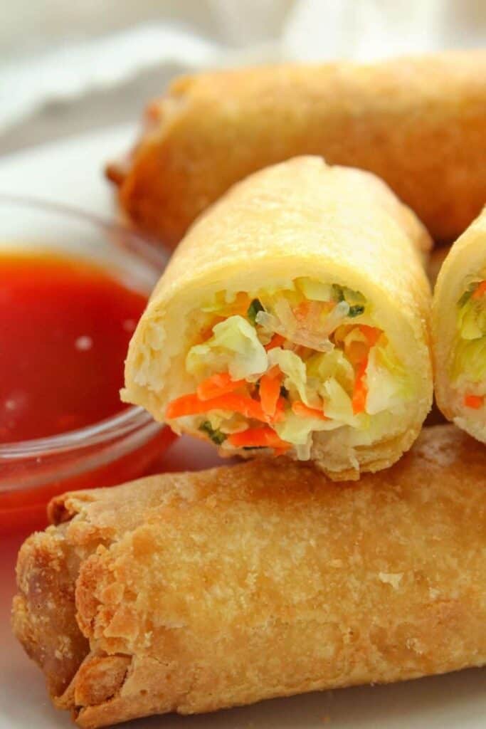 Closeup of frozen egg rolls on a white plate with one cut open and sauce in a small bowl on the side