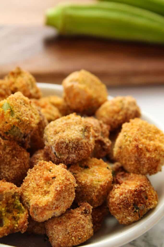 Air fryer okra in a small white bowl with fresh okra behind them.