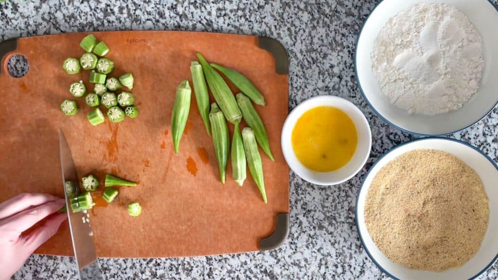 Fresh okra being cut into slices next to bowls of flour, egg, and cornmeal.