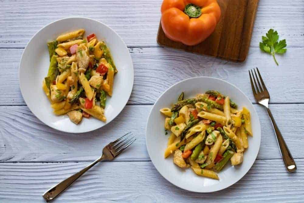 Two white plates of chipotle chicken pasta from overhead wit forks and an orange sweet pepper in the background