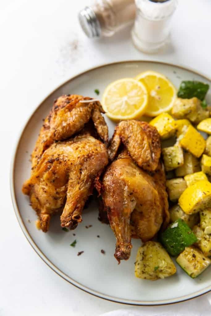 Air Fryer Cornish Hens cut in half next to each other on its side and served with lemons and zucchini and squash