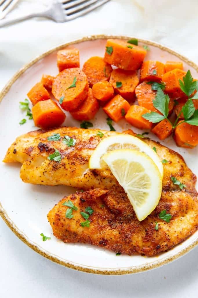 Tilapia on a white plate with lemon wedges and chopped carrots