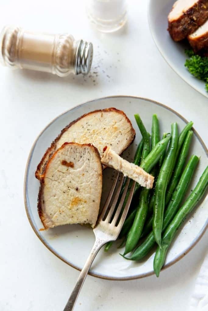 Closeup of pork loin served on a white plate with green beans with a piece cut on a fork