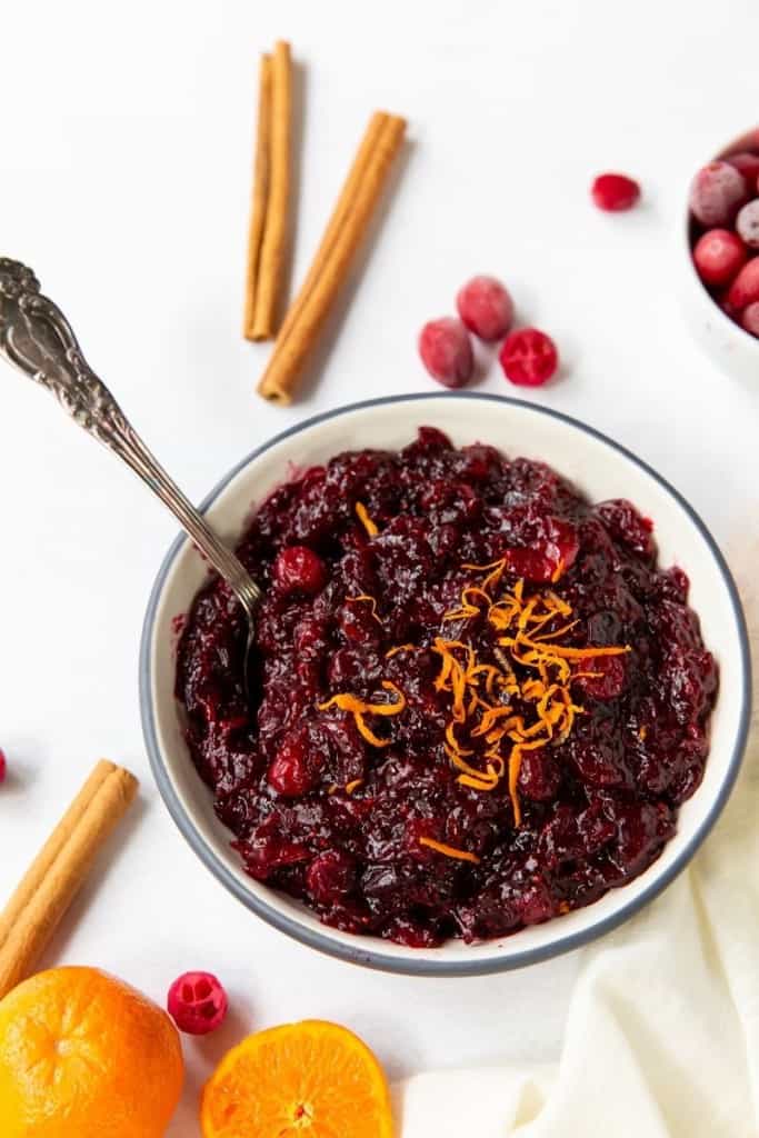 Bowl of cranberry sauce with orange zest on top with cinnamon sticks and oranges around it