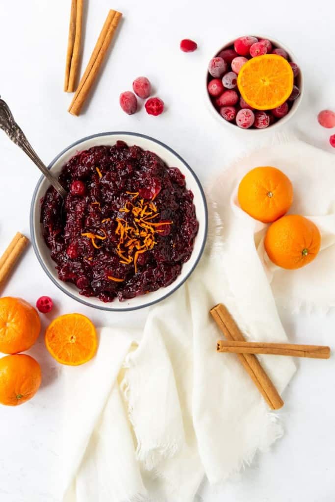 cranberry sauce in a bowl with a spoon surrounded by oranges ad cinnamon sticks