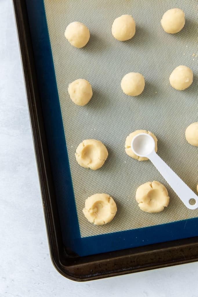 Cookies being pressed down in the center on a baking sheet with a teaspoon
