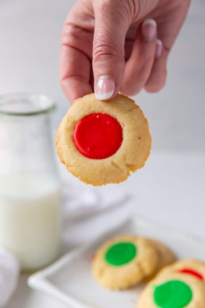 Holding a red iced thumbprint cookie in hand with a plate of cookies and milk in background