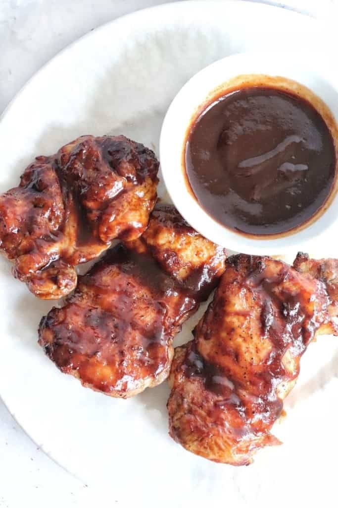 Three BBQ Chicken thighs on a white plate with a little bowl of barbecue sauce