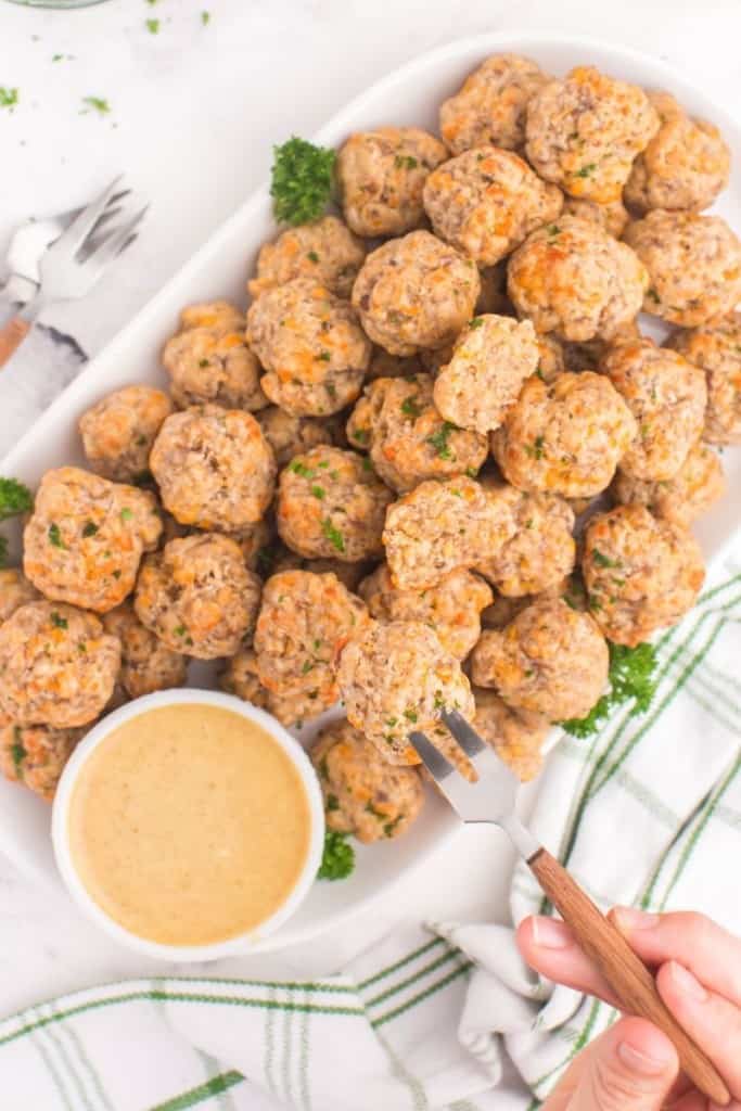 Overhead of sausage balls on a serving platter with honey mustard and serving fork with hand taking one