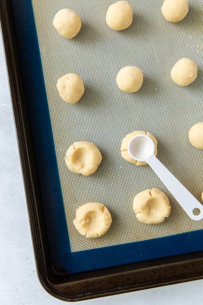 The back of a measuring spoon pressing into the thumbprint cookie recipe with icing to make a dent for the icing.