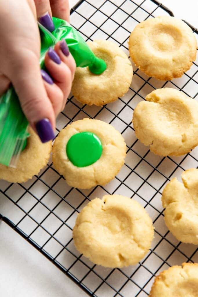 A hand icing the thumbprint cookies green on a wire rack.