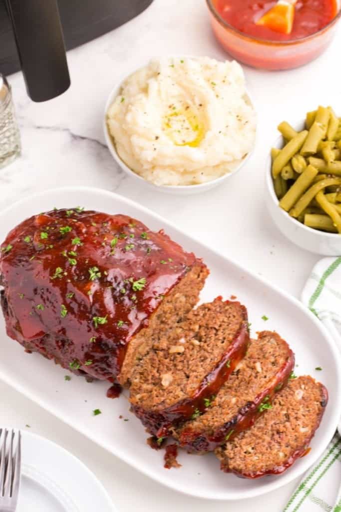 Sliced air fryer meatloaf on a serving tray with mashed potatoes and green beans in bowls in background