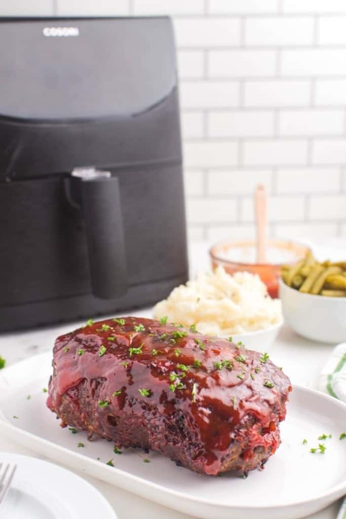 Full meatloaf on a white serving plate in front of an air fryer
