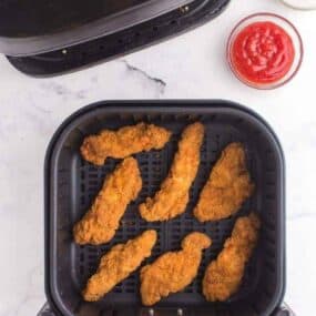 Cooked Frozen Chicken Tenders in the air fryer basket with a side of ketchup up top