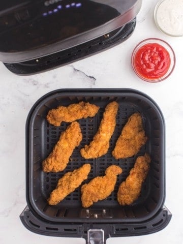 Cooked Frozen Chicken Tenders in the air fryer basket with a side of ketchup up top