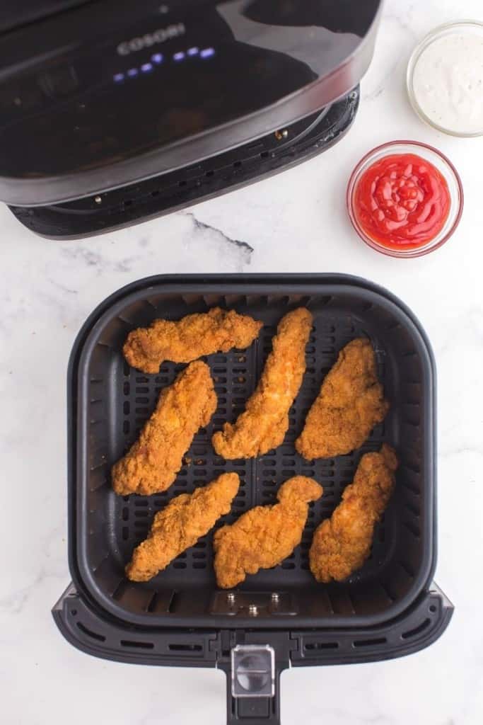 Cooked Frozen Chicken Tenders in the air fryer basket with a side of ketchup up top