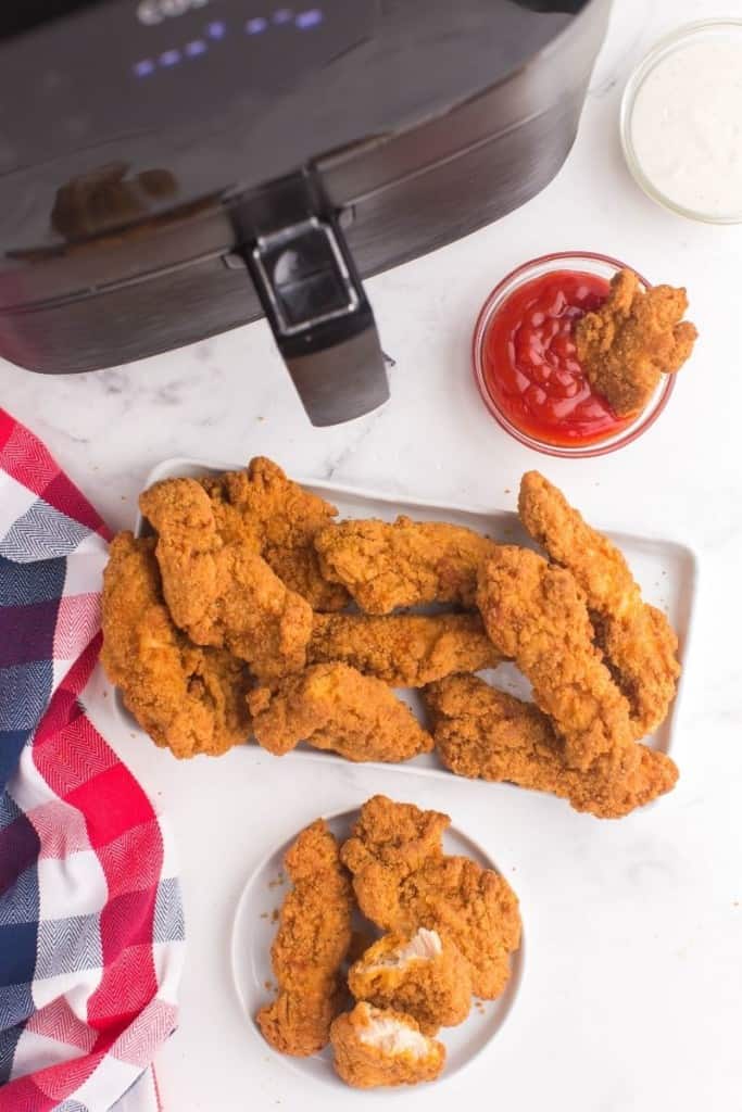 Air Fryer Frozen Chicken Tenders on a rectangular platter and a circular plate with a little bowl of ketchup next to an air fryer