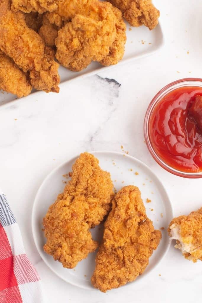 Chicken tenders on a white plate with ketchup in a small bowl to the side