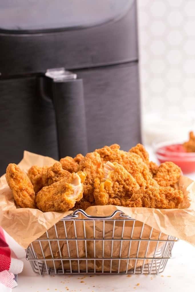 Frozen chicken fingers in a wired serving basket with parchment paper in front of an air fryer