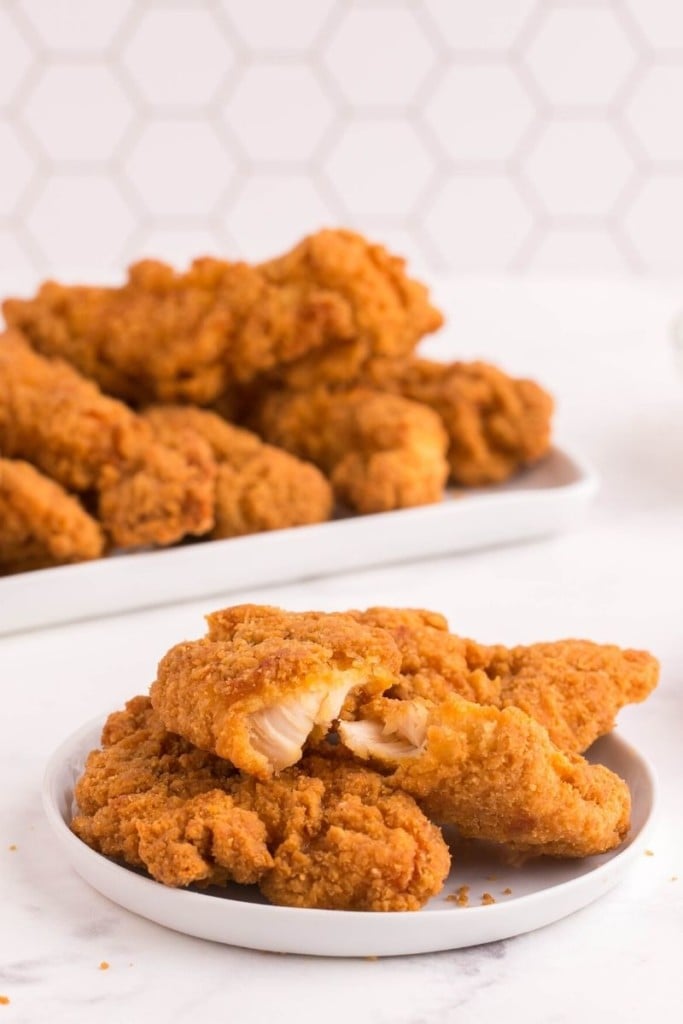 Air Fryer Frozen Chicken Tenders stacked on a white plate with a serving plate of them in the background