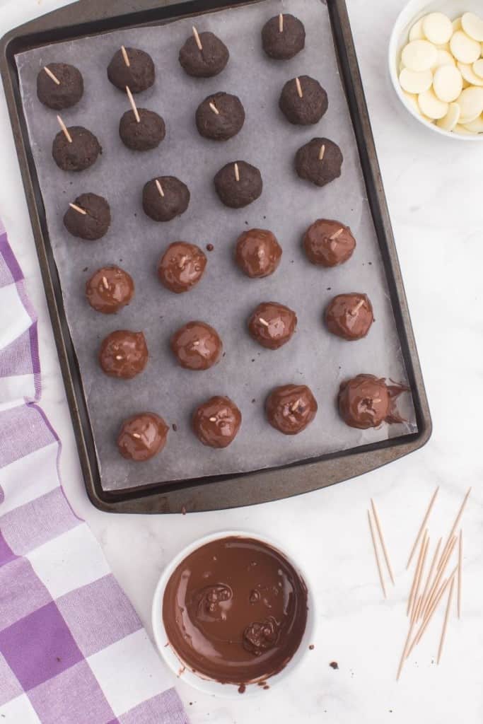 Oreo truffles with toothpicks in them and half of them covered in chocolate on a baking sheet and dipping sauce next to it