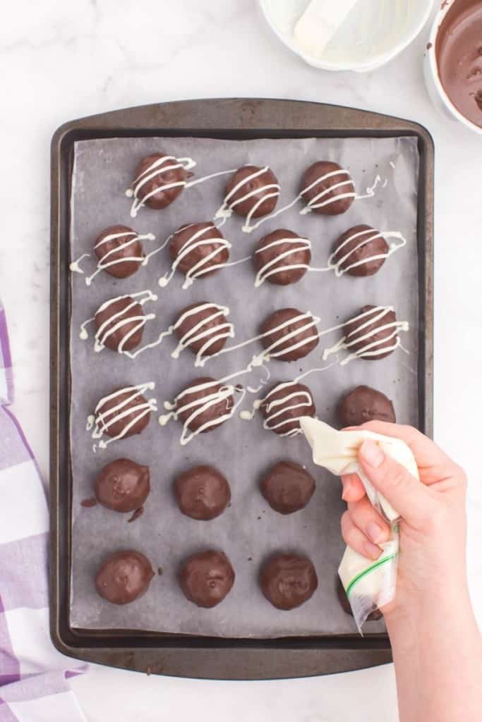 Oreo balls being piped with white chocolate on a baking sheet
