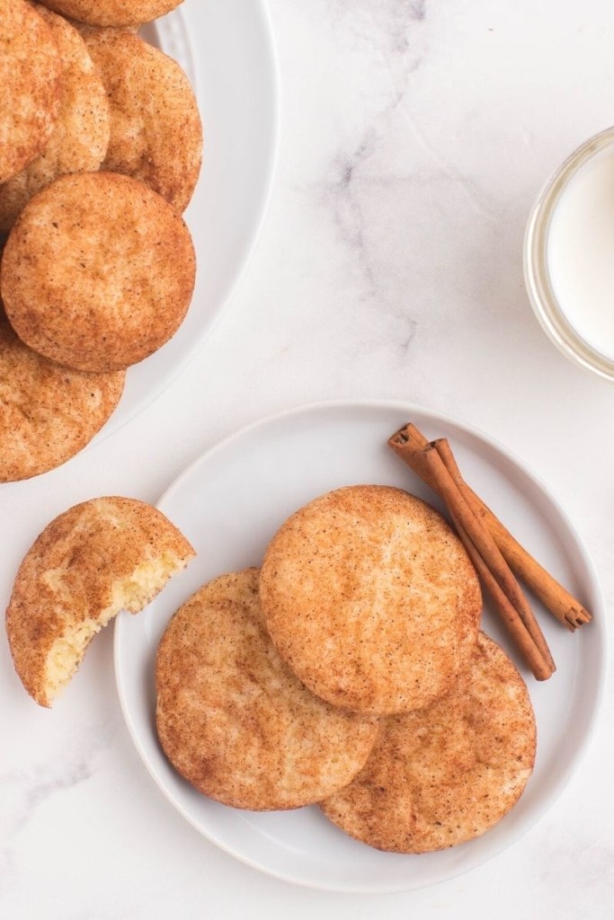 Snickerdoodles on a white round plate with cinnamon sticks and a serving plate of cookies to the side